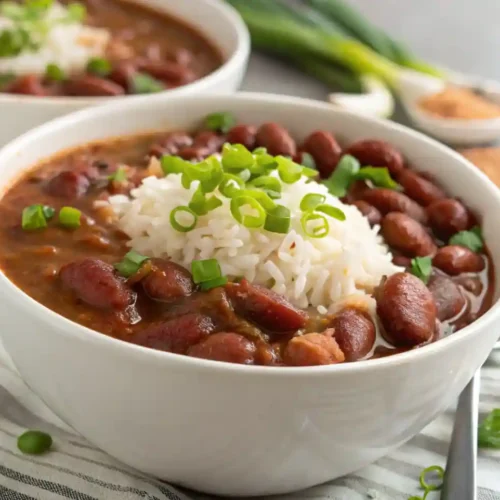 A bowl of authentic New Orleans-style Red Beans and Rice, garnished with chopped green onions, served in a white bowl with fluffy white rice on top. A second bowl and fresh ingredients like green onions and spices are visible in the background.